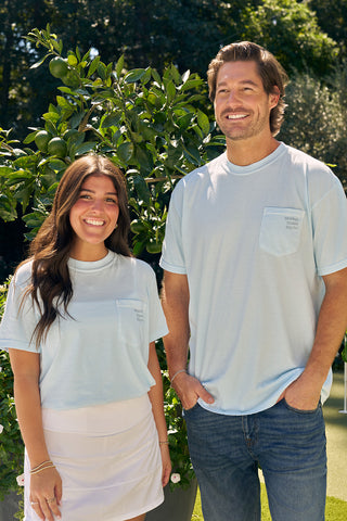A smiling woman in a white skirt and a man in jeans stand outdoors in front of leafy plants, both wearing light blue SDS Nashville '12 South' Tees with chest pockets. The woman has long brown hair; the man has short brown hair.