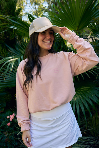A person wearing an SDS Nashville 12 South pink crewneck sweatshirt, a white skirt, and a beige cap smiles in front of lush green foliage.