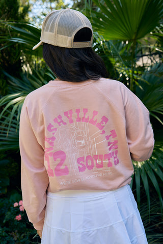 A person stands outdoors in a SDS Nashville 12 South Crewneck Sweatshirt, featuring Nashville, TN 12 South on the back. A beige cap tops the look, while lush green palm leaves frame the scene.