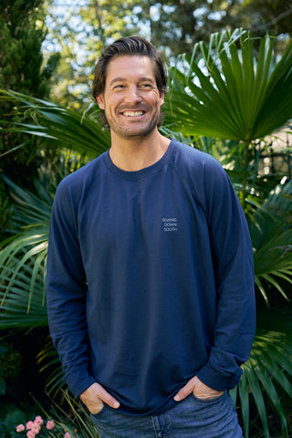A man smiles in an SDS King Street Crewneck Sweatshirt, Indigo, and jeans, surrounded by vibrant green plants. The shirt displays Sewing Down South, and tall leafy foliage provides a scene evoking the charm of a Charleston storefront.