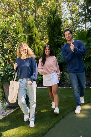 Three people stroll along a grassy path. On the left, someone with curly hair and a blue sweater carries a tote bag. The middle person sports a pink sweater and white skirt, while the right wears jeans and an SDS Nashville 12 South crewneck sweatshirt in blue.