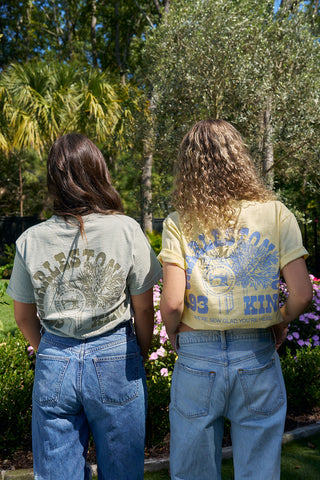 Two women with long hair stand outdoors, backs to the camera, wearing SDS 'King Street' Flagship Tees and blue jeans in a sunlit garden filled with flowers, green grass, and palm trees—an ideal Sewing Down South moment.
