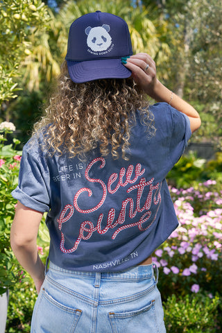 A person with curly hair wears a blue ‘Life is Better in Sew Country’ unisex tee and a navy cap with a panda logo, standing outdoors in a garden with green foliage and pink flowers.