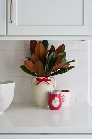 A white vase with a red ribbon and magnolia leaves sits on a white kitchen counter beside a Sewing Down South Santa Wine Tumbler and a white mug with red lettering, set against a backdrop of white tile backsplash and cabinetry.