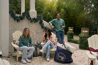 Two women in Nutcracker Long Sleeve Tees smile outdoors on Adirondack chairs with a man standing behind them, all holding drinks. A large navy bag filled with greenery sits near their decorated patio.
