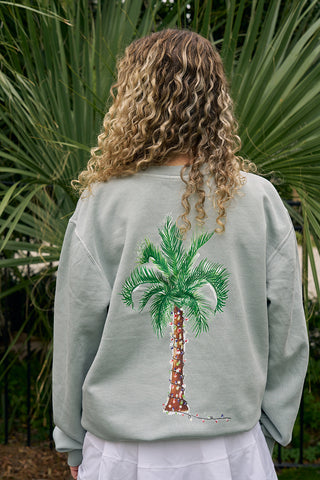 A person with curly blonde hair stands outdoors before large green palm leaves, wearing the Holiday Palmetto Crewneck—a coastal holiday sweatshirt featuring a palm tree graphic with string lights.