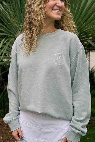 A person with curly blonde hair smiles while wearing the Holiday Palmetto Crewneck in light green with white chest text over a white shirt. Palm fronds appear in the background.