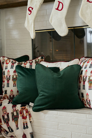 Two green Nutcracker Stripe Pillows in Spruce rest on a white brick fireplace. Above, cream knit stockings with red letters hang, completing the festive holiday setting with nutcracker charm.