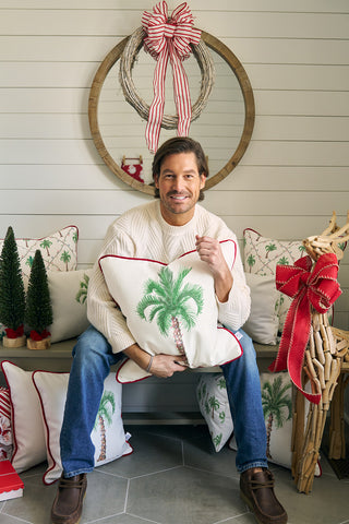 A man in a cream sweater and jeans sits on a bench holding an Oversized Holiday Palmetto Pillow, surrounded by festive Christmas trees, cozy pillows, and a wicker reindeer, with a round mirror and holiday wreath behind him.