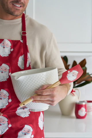 A smiling person in a Santa apron holds the BPA-free silicone Santa Spatula and a white mixing bowl, standing in a festive kitchen—making the Santa Spatula an ideal holiday baking gift.