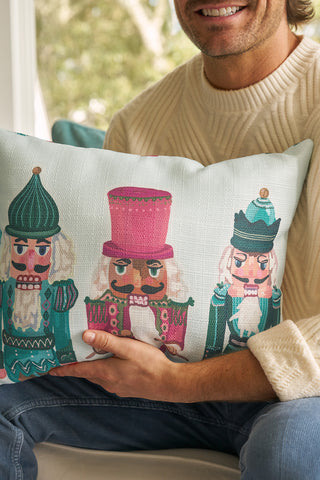 A person in a cream sweater holds the Pastel Nutcracker Lumbar Pillow, which features three colorful nutcracker designs, while sitting indoors as sunlight streams through a window in the background.