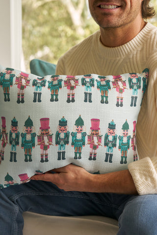 A man in a cream sweater holds the Pastel Nutcracker Lumbar Pillow, which features festive, colorful nutcrackers in green and pink uniforms. He sits indoors with the holiday pillow, his face partially out of frame.