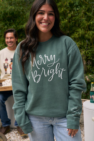 A woman with long dark hair smiles at the camera, wearing the Merry + Bright Crewneck. She stands outdoors with another person and greenery in the background.
