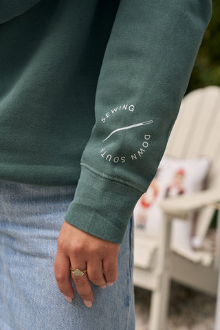 A close-up of a person wearing the Merry + Bright Crewneck—a cozy cotton blend green holiday sweatshirt with white text and a needle graphic reading SEWING DOWN SOUTH on the sleeve—paired with light blue jeans and a gold ring.