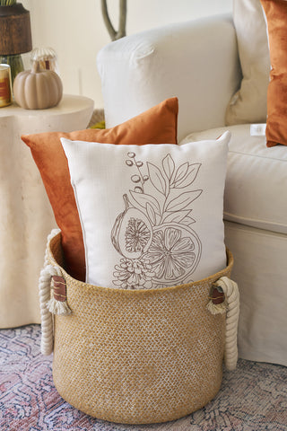 A woven basket with rope handles holds two pillows—one solid burnt orange velvet accent pillow and one Holiday Harvest Lumbar Pillow, featuring a line drawing of fruit and leaves. The basket is on a patterned rug beside a white armchair.