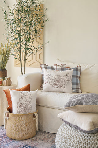 Neutral-toned living room with a white sofa, Holiday Harvest Plaid pillow, patterned throw pillows, woven basket with pillows, knit pouf, stacked cushions, leafy plant, and wall art for a Southern-inspired look.