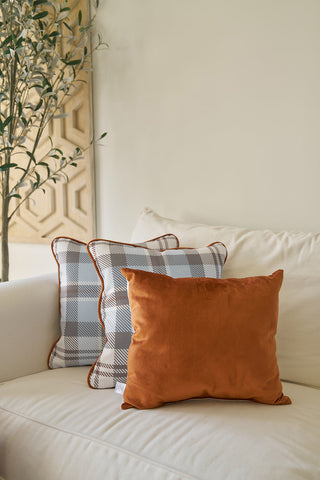 A Holiday Harvest Lumbar Pillow sits on a cream-colored couch beside a blue and white checkered pillow with brown piping. In the background, a leafy plant and wooden wall panel can be seen.