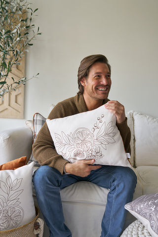 A smiling man with brown hair sits on a white couch, holding the Holiday Harvest Lumbar Pillow featuring leafy and citrus fruit designs. Autumnal decor pillows surround him, with a plant and wall decor in the background.