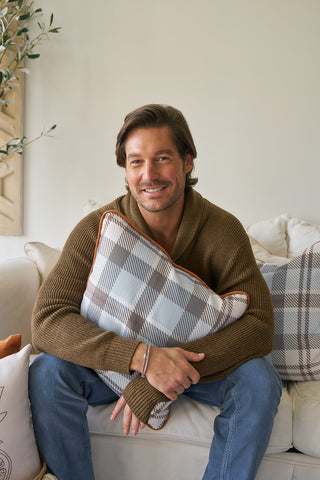 A man with brown hair and a beard smiles on a white couch, hugging a Holiday Harvest Plaid pillow. He wears a brown sweater and blue jeans, surrounded by cozy decor that adds to the inviting Southern style of the space.