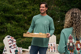 A man in a green Nutcracker Long Sleeve Tee holds a wooden tray with smores supplies outdoors, smiling at a woman with curly hair. White chairs with festive nutcracker-themed pillows and blankets are in the background.