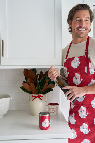 A man in a festive red Santa apron smiles while opening wine in a holiday-decorated kitchen featuring a Santa mug, the Santa Wine Tumbler, and a vase filled with greenery wrapped in a red ribbon.