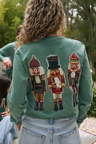 Someone with curly hair wears a green Nutcracker Long Sleeve Tee, featuring three colorful nutcrackers on the back and Sewing Down South near the hem—a festive, holiday-themed shirt.