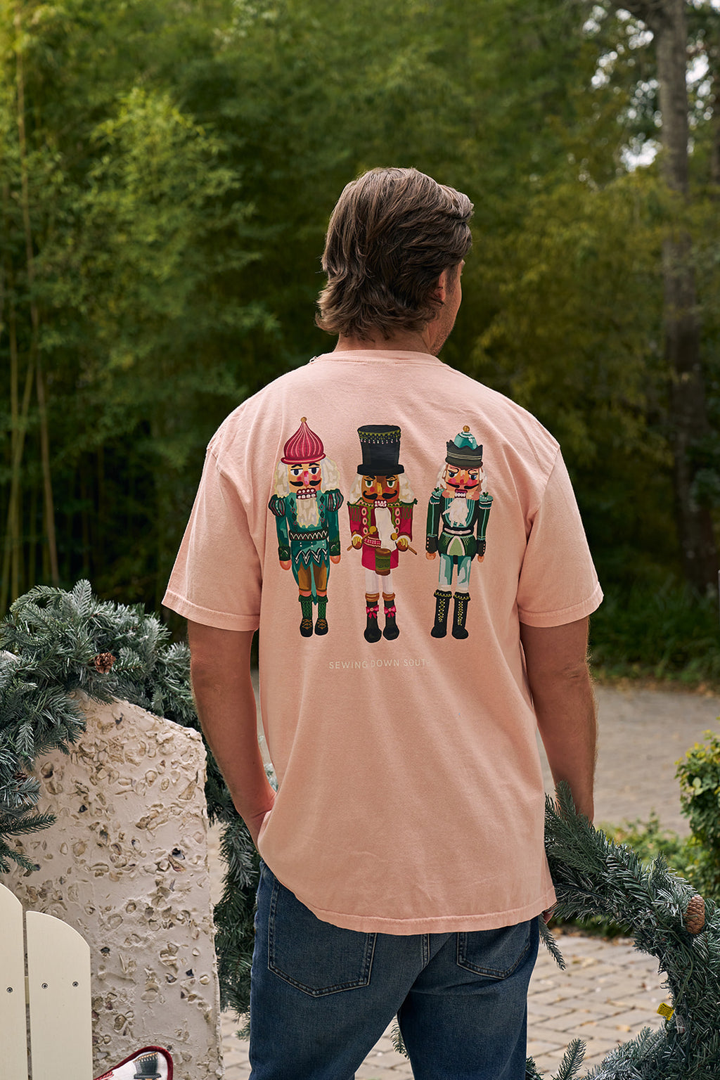 A person wearing the Pastel Nutcracker T-shirt, featuring three colorful nutcracker designs on the back, stands outdoors near a decorated garland with greenery in the background—a festive holiday apparel choice.