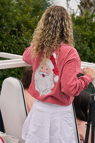 A person with curly blonde hair wears a Santa Long Sleeve Tee—a festive red unisex shirt—paired with a white skirt, standing beside a white vehicle outdoors near greenery.