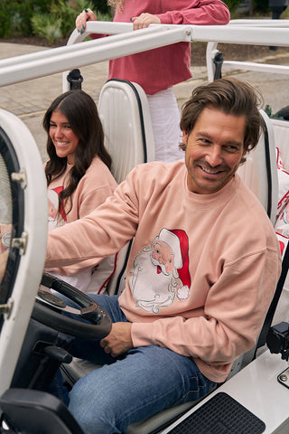 A smiling man and woman sit in a white open vehicle, both wearing light pink Santa Crewneck sweatshirts. Another person stands behind the car, partially visible, creating a cheerful and casual scene.