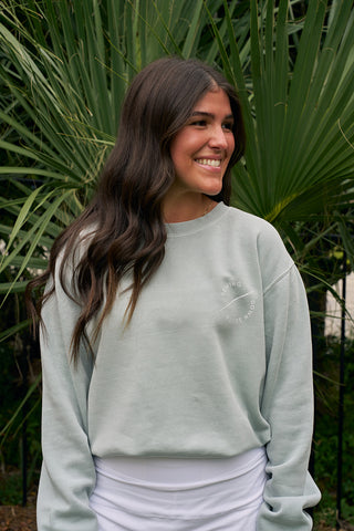 A woman with long, wavy brown hair smiles outdoors in front of lush palm leaves, wearing the Holiday Palmetto Crewneck sweatshirt in light gray and a white skirt.