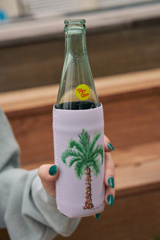 A person with teal-painted nails holds a glass Topo Chico bottle in a light purple Holiday Palmetto Koozie, 4 PACK (Skinny or Standard), decorated with a palm tree illustration.