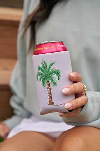 A person in a light sweater holds a can of strawberry lemon drink in the Holiday Palmetto Koozie, 4 PACK (Skinny or Standard)—a light purple neoprene koozie with a palm tree design. Only their hand and partial arm are visible.