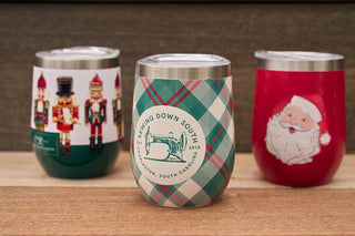 Three holiday-themed insulated cups are shown on a wooden surface. The center cup is the Holiday Plaid Wine Tumbler with the Sewing Down South logo; the left has nutcrackers, and the right features a Santa Claus illustration.