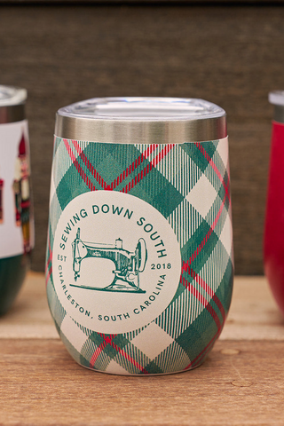 The Holiday Plaid Wine Tumbler, decorated with a vintage sewing machine and SEWING DOWN SOUTH EST 2018 CHARLESTON, SOUTH CAROLINA, sits on a wooden surface among other colorful insulated tumblers.