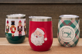 The Santa Wine Tumbler, shown on a wooden surface, is red with a jolly Santa face. Two more tumblers behind it display nutcracker designs and a green plaid pattern with text—perfect for festive holiday toasts.