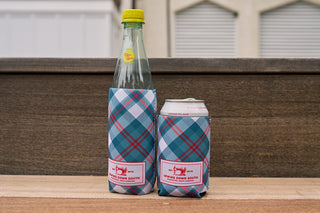 Two drinks—a glass bottle and a can—each in a Holiday Plaid Koozie, 4 PACK (Skinny or Standard), featuring the Sewing Down South logo, rest on a wooden surface with a blurred background.