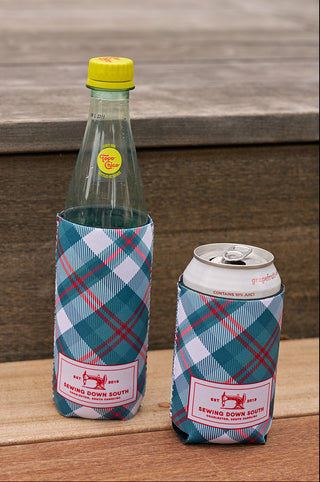 Two drinks—one in a glass bottle with a yellow cap and label, the other in a can—sit on wooden steps, each wrapped in a Holiday Plaid Koozie, 4 PACK (Skinny or Standard) labeled SWING DOWN SOUTH for a festive touch.
