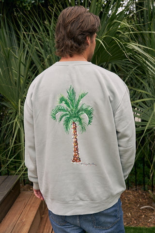 A person with light brown hair stands outdoors before tall greenery, wearing the Holiday Palmetto Crewneck which features a palm tree graphic on the back.