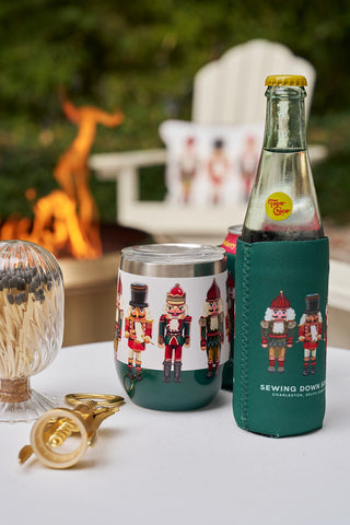 A set of Nutcracker Koozies (4 pack, skinny or standard), featuring festive nutcracker designs, sits on a white outdoor table alongside matching holiday décor.