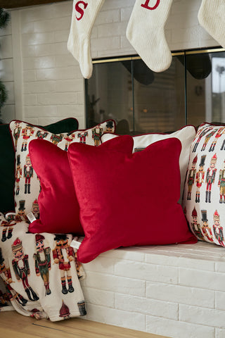 Two Scarlet Velvet Pillows rest on a white bench with a Nutcracker Stripe Pillow in Scarlet and a blanket. Above, white Christmas stockings hang from the mantel, showing partial letters S and D.