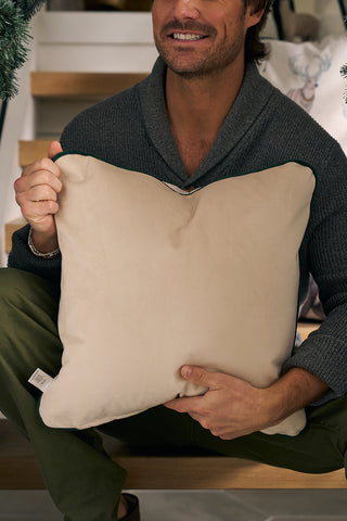A man sits on stairs, smiling and holding the Holiday Reindeer Pillow in front of his chest. He wears a dark gray sweater with green pants, creating a cozy indoor scene that highlights the pillows festive charm.
