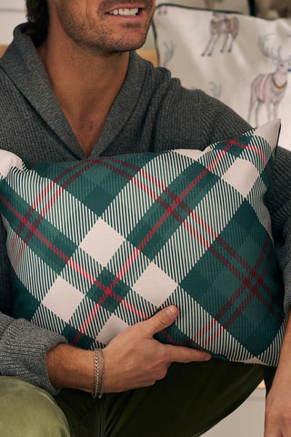 A man in a gray sweater holds a Green & Red Plaid Lumbar Pillow by Sewing Down South. Behind him is a decorative pillow with a reindeer illustration; his face is partially visible.
