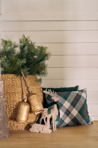 A woven basket with pine branches and gold bells sits next to a Green & Red Plaid Lumbar Pillow, green pillows, and a tan decorative reindeer on a wooden floor against a white shiplap wall.