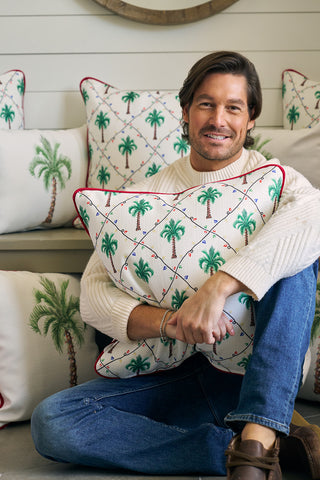 A man in a cream sweater and blue jeans sits cross-legged, smiling as he hugs an Oversized Holiday Palmetto Pillow featuring a palm tree design. More oversized palm tree pillows are stacked behind him.