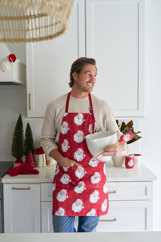 A man in a Santa Apron with an adjustable neck strap holds a white mixing bowl and whisk, smiling in a festive kitchen decorated with small Christmas trees and holiday décor on a white counter.