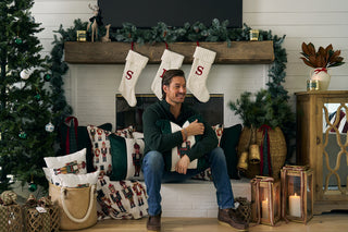 A man smiles before a festive fireplace with stockings, gifts, garlands, and pillows. The Nutcracker Stripe Pillow in Spruce and collectible nutcrackers bring extra charm to the cozy holiday scene.