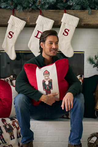 A man in a green sweater and jeans sits by the fireplace, holding a Nutcracker Stripe Pillow in Scarlet. Three white stockings with red letters hang above the mantel, which is beautifully adorned with pine garland.