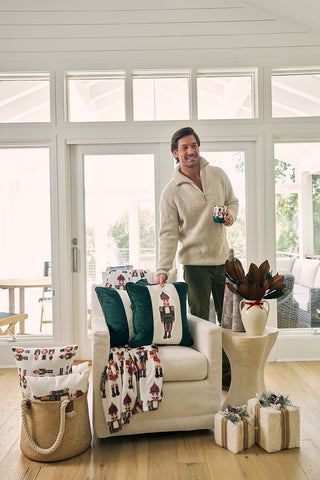 A smiling man in a cream sweater stands by a cozy armchair, topped with the Nutcracker Stripe Pillow in Spruce and a festive blanket. Wrapped gifts and a potted plant sit nearby in the sunlit living room with large windows.