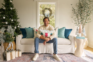 A smiling man sits on a white sofa holding the Pastel Nutcracker Lumbar Pillow, surrounded by festive decorations, a Christmas tree, wrapped gifts, and teal cushions in a bright living room.