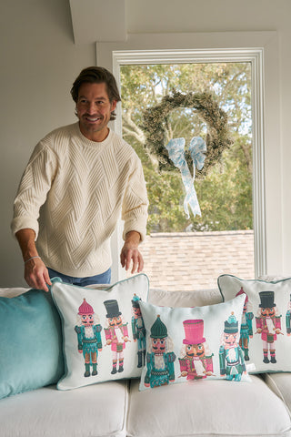 A man in a cream sweater smiles behind a couch decorated with the Pastel Nutcracker Pillow and other holiday pillows. A wreath with a blue ribbon hangs in the window behind him.